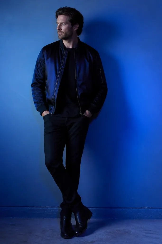 A man stands in front of a blue LED wall wash. He wears a black satin bomber jacket over a matte black tee, black jeans, and black boots. The satin catches the blue light with a smooth sheen.