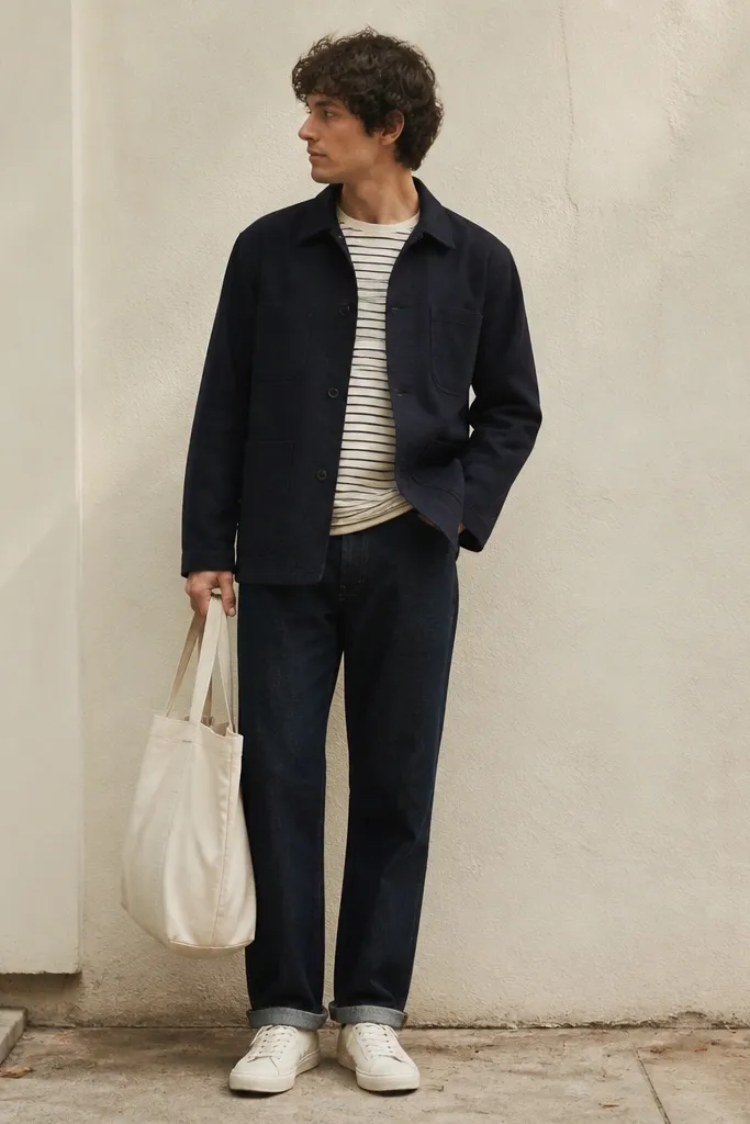 A navy chore coat worn over a striped tee, paired with dark raw denim and white low-top sneakers; the outfit looks relaxed but structured, with a canvas tote bag.