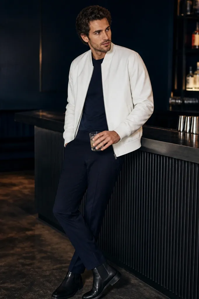 A man wearing a white bomber jacket with ribbed cuffs over a dark tee. He has navy trousers and black Chelsea boots. The background shows a dim lounge with blue lighting and a bar counter.