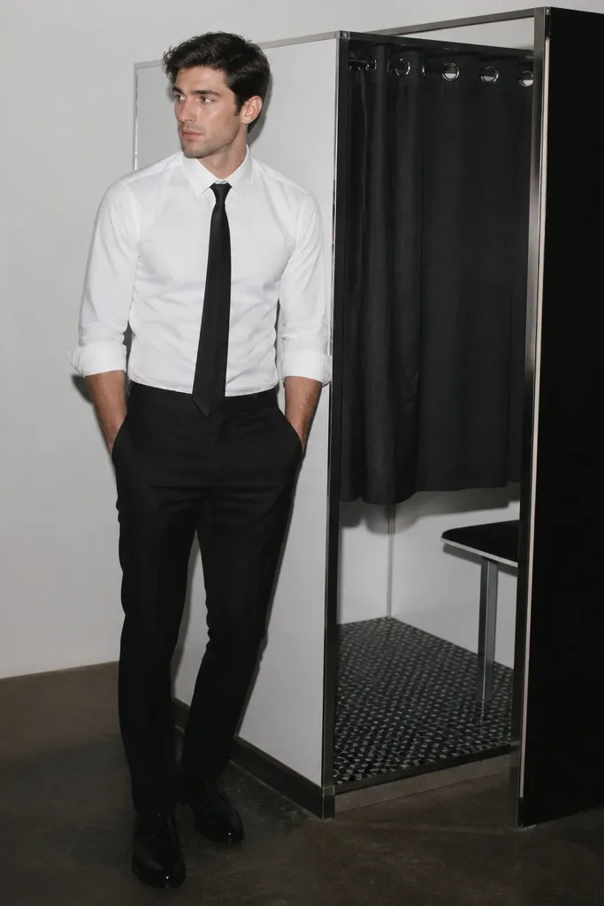 A man wearing a crisp white button-down with sleeves rolled slightly and a black skinny tie. He pairs it with black trousers and black dress shoes. The background shows a photo booth with bright flash lighting.