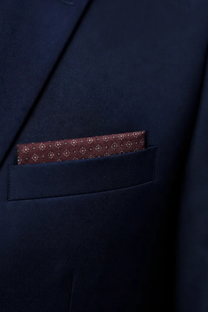 A jacket breast pocket is shown with a neatly folded pocket square insert. The insert is removable and sits flush in the pocket. The jacket is dark navy, and the pocket square has a muted burgundy pattern.