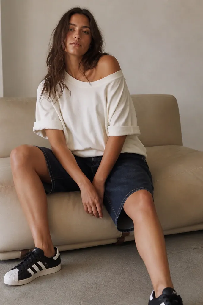A woman wearing black Adidas Superstar sneakers, dark indigo denim jorts with a slightly longer inseam, and an oversized cream T-shirt. The tee drapes off one shoulder slightly, sleeves are cuffed, and the jorts have minimal distressing.