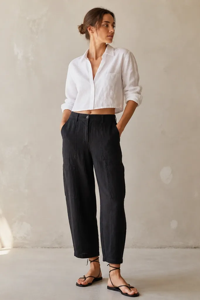 Black linen pants with storage seam detailing at the side. A white cropped button-up sits above the waistband, sleeves rolled. Black strappy sandals wrap around the ankle.
