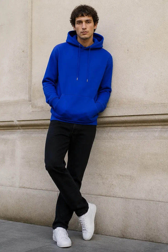 A bright cobalt blue hoodie worn over black jeans with a clean straight leg. The hoodie strings hang down, and the person wears white leather sneakers. The background is an outdoor street wall, making the blue pop.