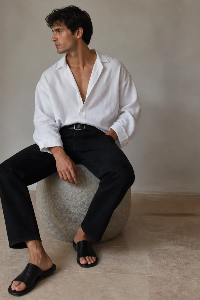 A man wears black jeans with an oversized white linen shirt. It's open at the top with one button fastened, sleeves are loose, and he wears black leather slides and a black belt.