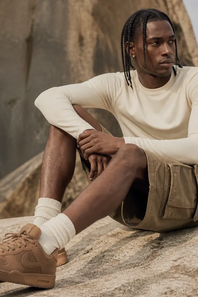 Brown Travis Scott-style sneakers in a lighter milk-chocolate tone with a smooth upper and textured details, photographed outdoors. The outfit shows tan cargo shorts, a fitted long-sleeve tee in warm white, and crew socks peeking above the shoe collar.