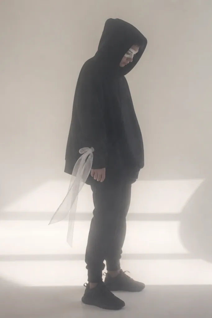 A person in an oversized black hoodie with the hood up, wearing black joggers and black sneakers. They have a simple white face paint around the eyes and a translucent white fabric strip tied at the wrist. The photo shows a ghostly, soft silhouette.