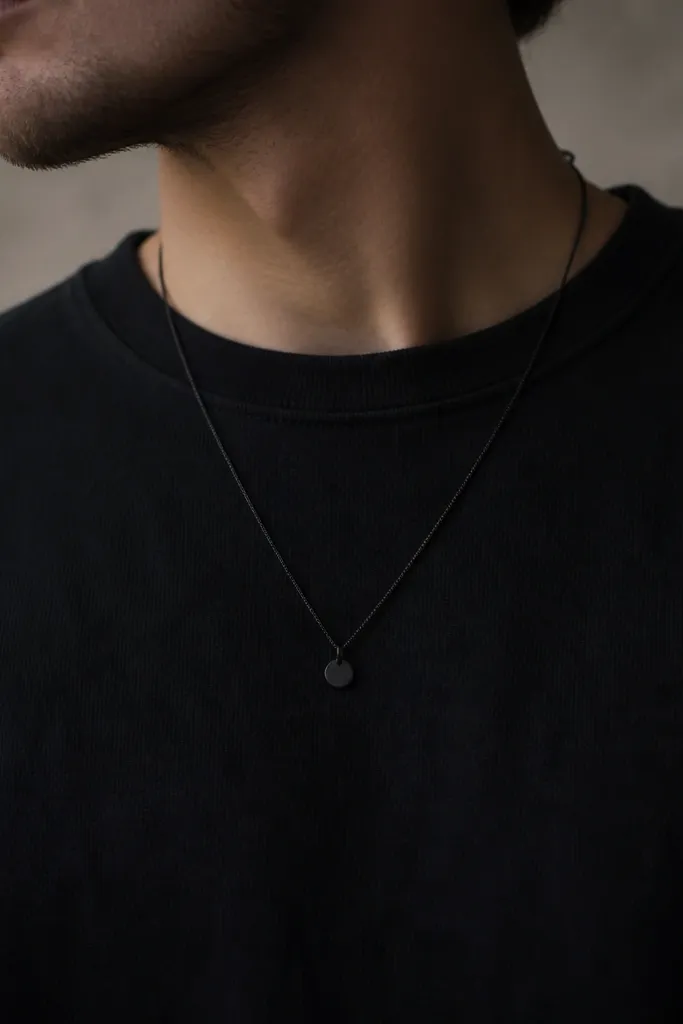 A thin black chain necklace with a small matte pendant resting on a black crewneck; the pendant is small and dark, not shiny.
