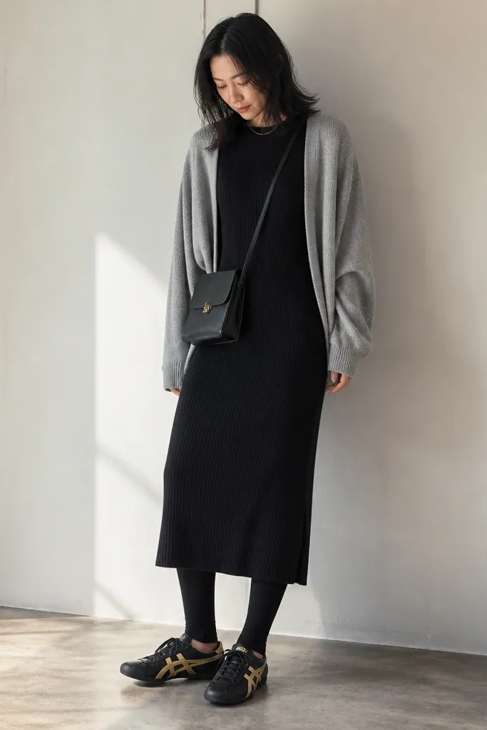 A black ribbed knit midi dress with a light gray cardigan draped open. The model wears black leggings underneath and black-and-gold Onitsuka Tiger sneakers. A small crossbody bag hangs at hip level.