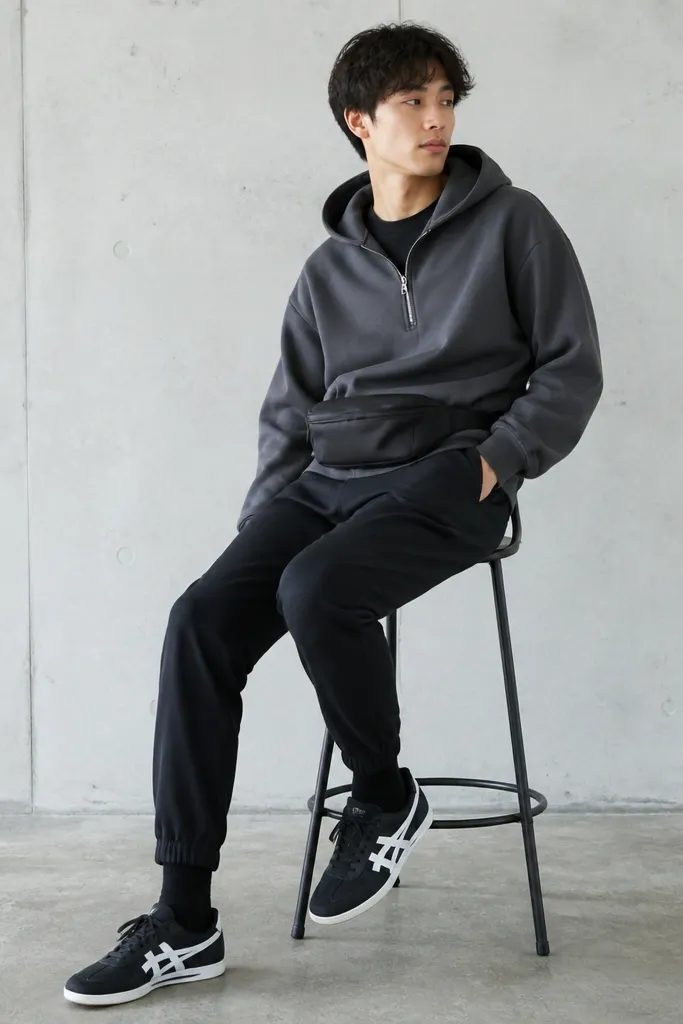 A model in a charcoal gray pullover hoodie, black tapered jogger pants with elastic cuff detail, black ankle socks, and Onitsuka Tiger mid runners in black and white. The jacket is unzipped slightly at the front, with a simple belt bag across the waist.
