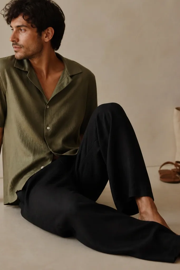 Black linen pants with an olive camp-collar short-sleeve shirt worn buttoned halfway. A pair of brown leather sandals and a simple canvas tote sit in the background.