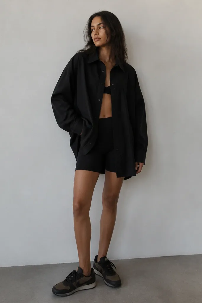 An oversized black button-up shirt worn open over black cycling shorts. Black Travis Scott shoes are visible with a relaxed stance.