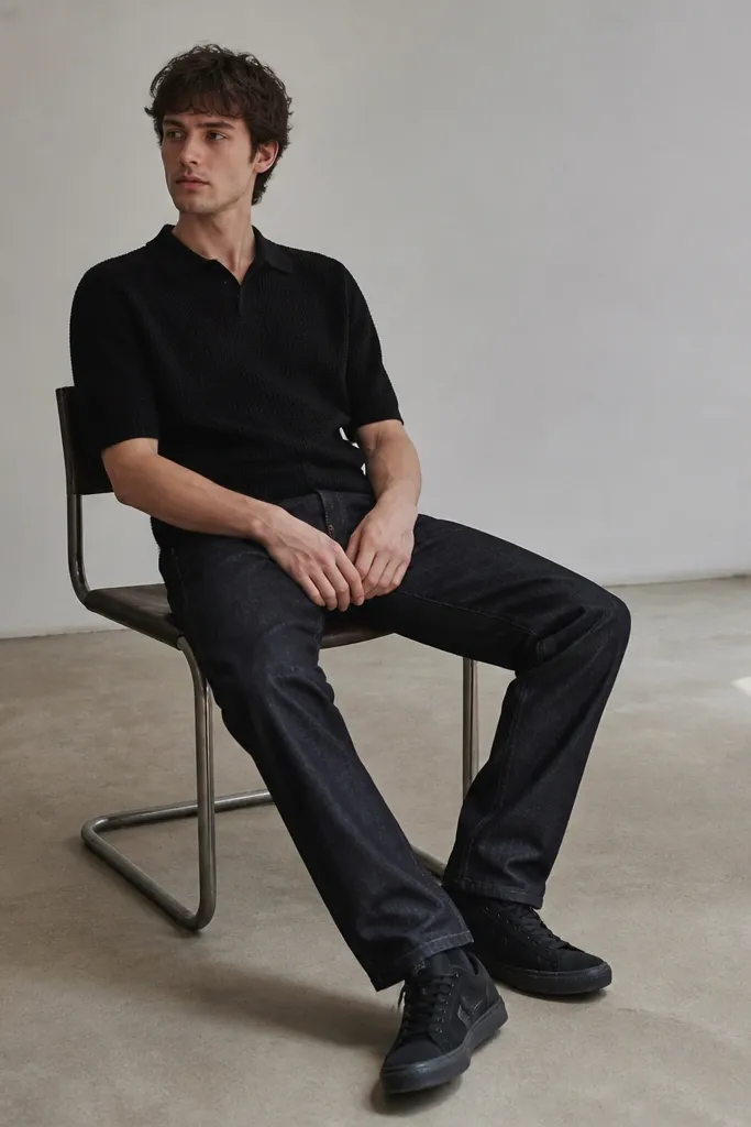 A black knit polo shirt with a short collar worn over dark wash jeans. Black Travis Scott sneakers are visible with a clean, slightly tapered leg.