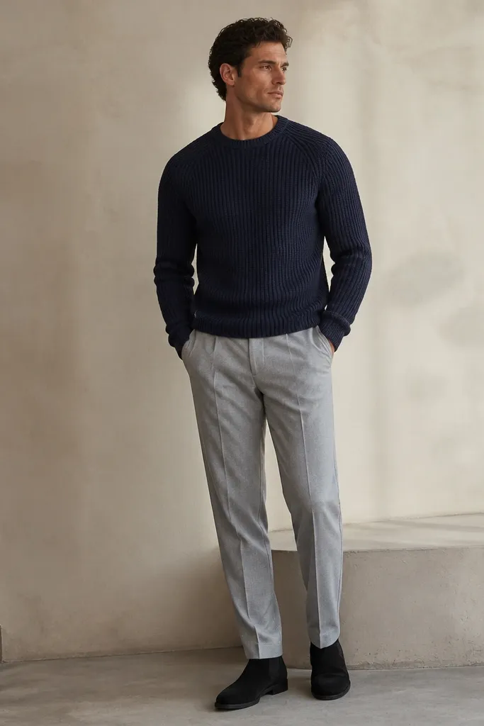 Black suede chelsea boots with light gray trousers, a navy knit sweater, and a simple outfit with no jacket. The trousers have a soft break and the sweater looks thick and textured.