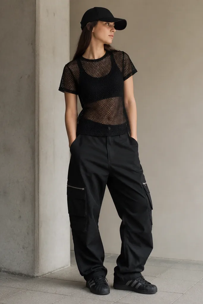 A black mesh short-sleeve top layered over a black tank, black cargo pants with zip pockets, black Superstar sneakers, and a black baseball cap.