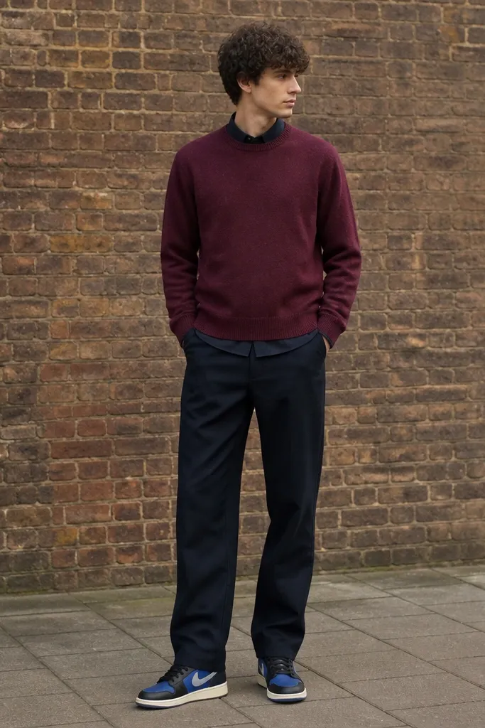 A model wears a burgundy crewneck sweater over a dark shirt, paired with navy straight pants. The blue Travis Scott shoes stand out under the hem with a storage seam near the heel. The background is a brick wall with overcast light.